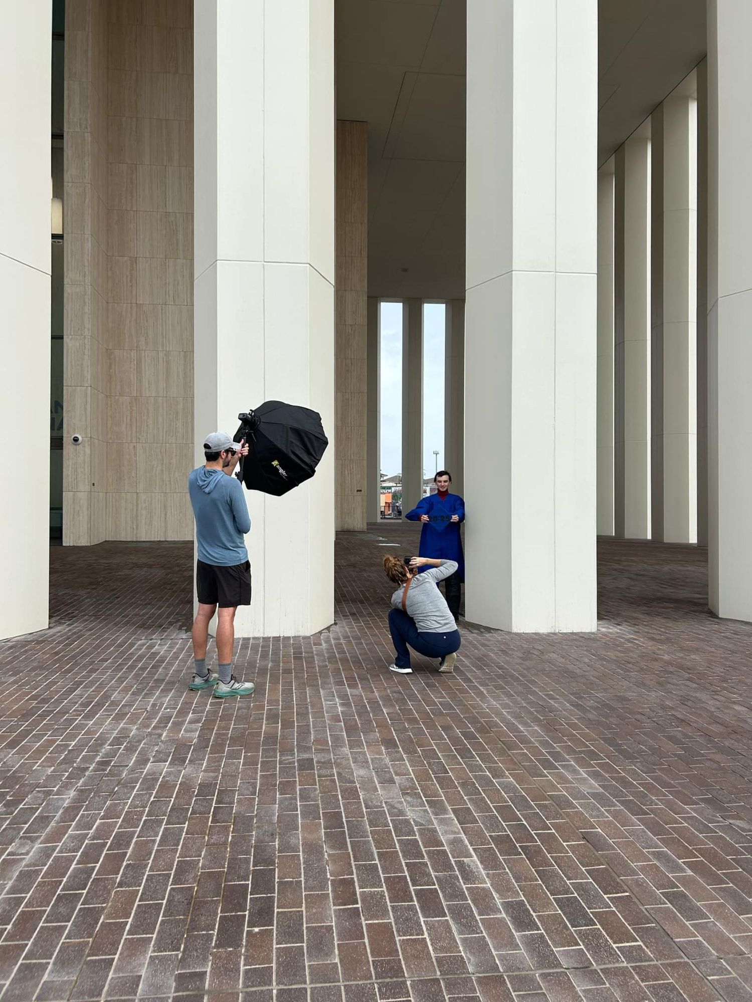 Photographer with umbrella taking pictures between large white columns in a modern architectural space with brick flooring.
