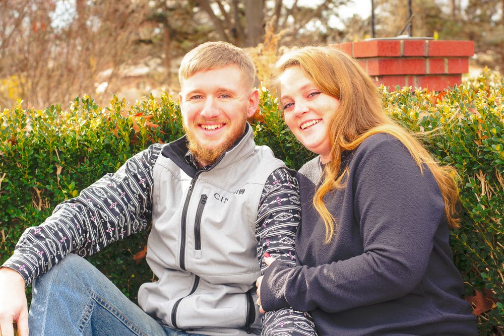 Smiling couple, man in gray vest with patterned shirt, woman in dark sweater, sitting outdoors amongst bushes.