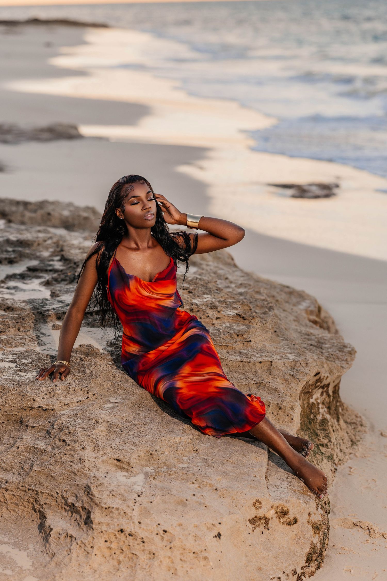 Woman in flowing dress poses dramatically on coastal rocks at sunset.