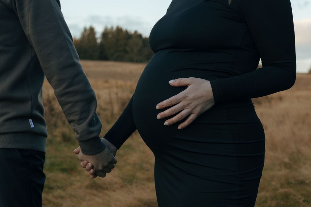 Close-up of two people holding hands while standing in autumn field, with expecting mother's round belly visible.