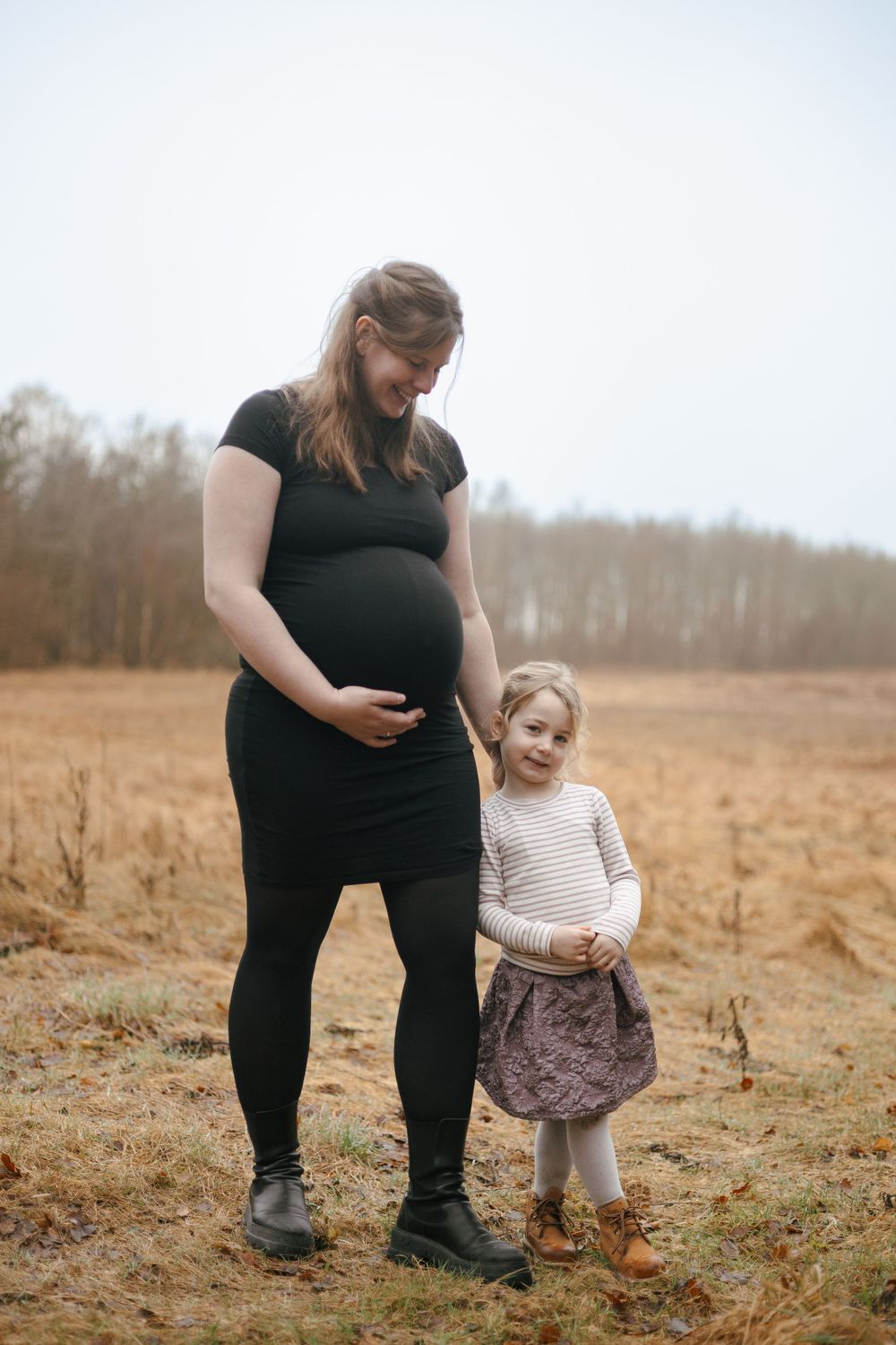 An expectant mother and young child stand together in a brown autumn field with bare trees in the background.