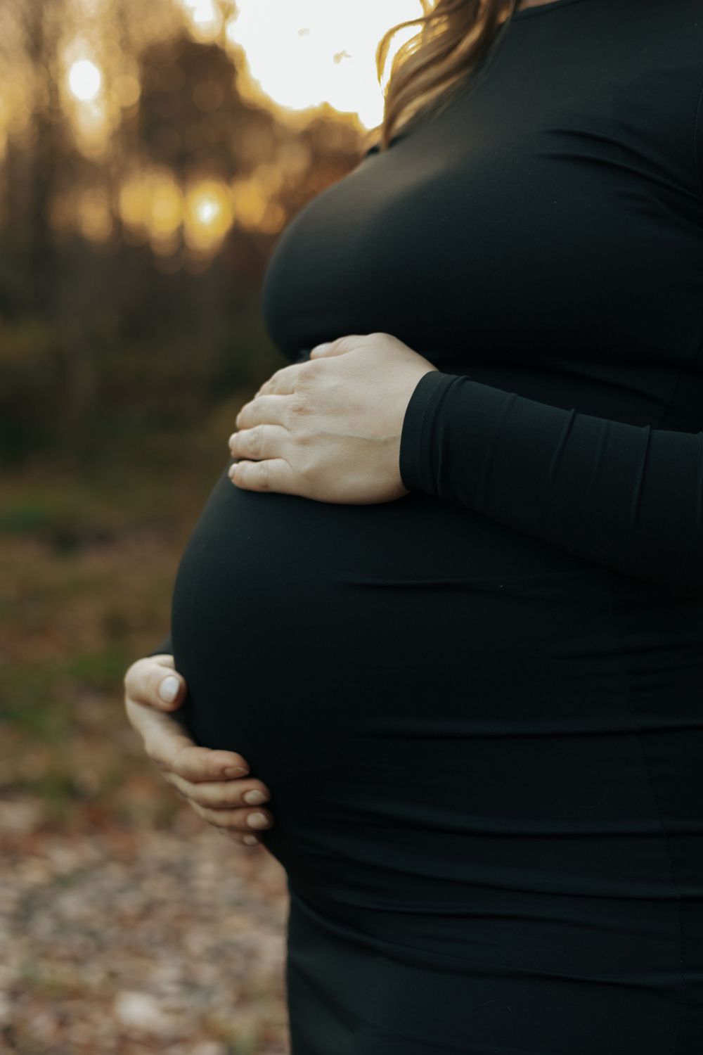 A pregnant woman in a black dress cradles her belly during sunset, creating a silhouette against golden bokeh lights.