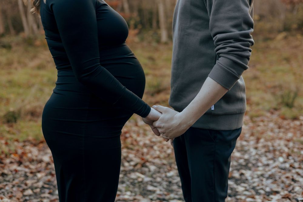 A couple holds hands tenderly during an outdoor maternity photoshoot with fall foliage in the background.