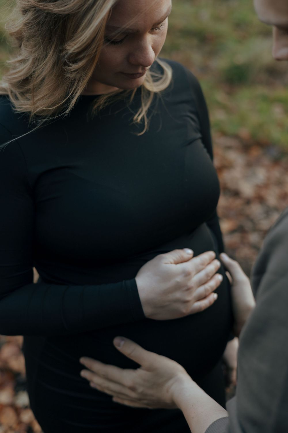 A woman in a black sweater holds her pregnant belly outdoors while hands reach to touch her bump on a fall day.