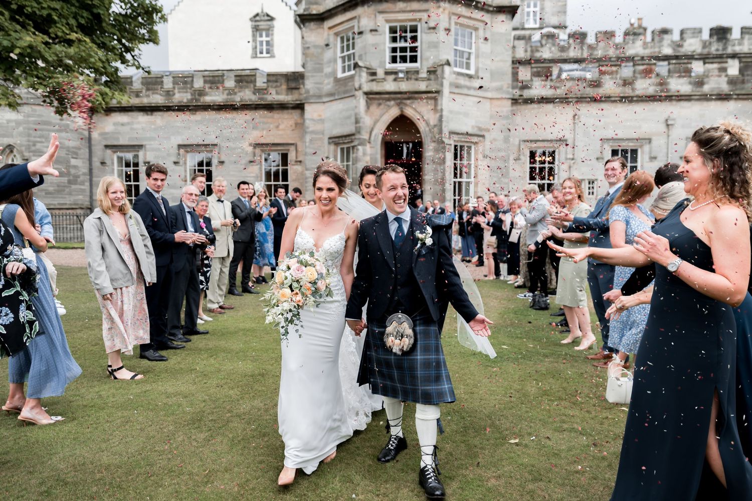 Newlyweds walk down grass path at a Scottish castle while wedding guests throw confetti in celebration.
