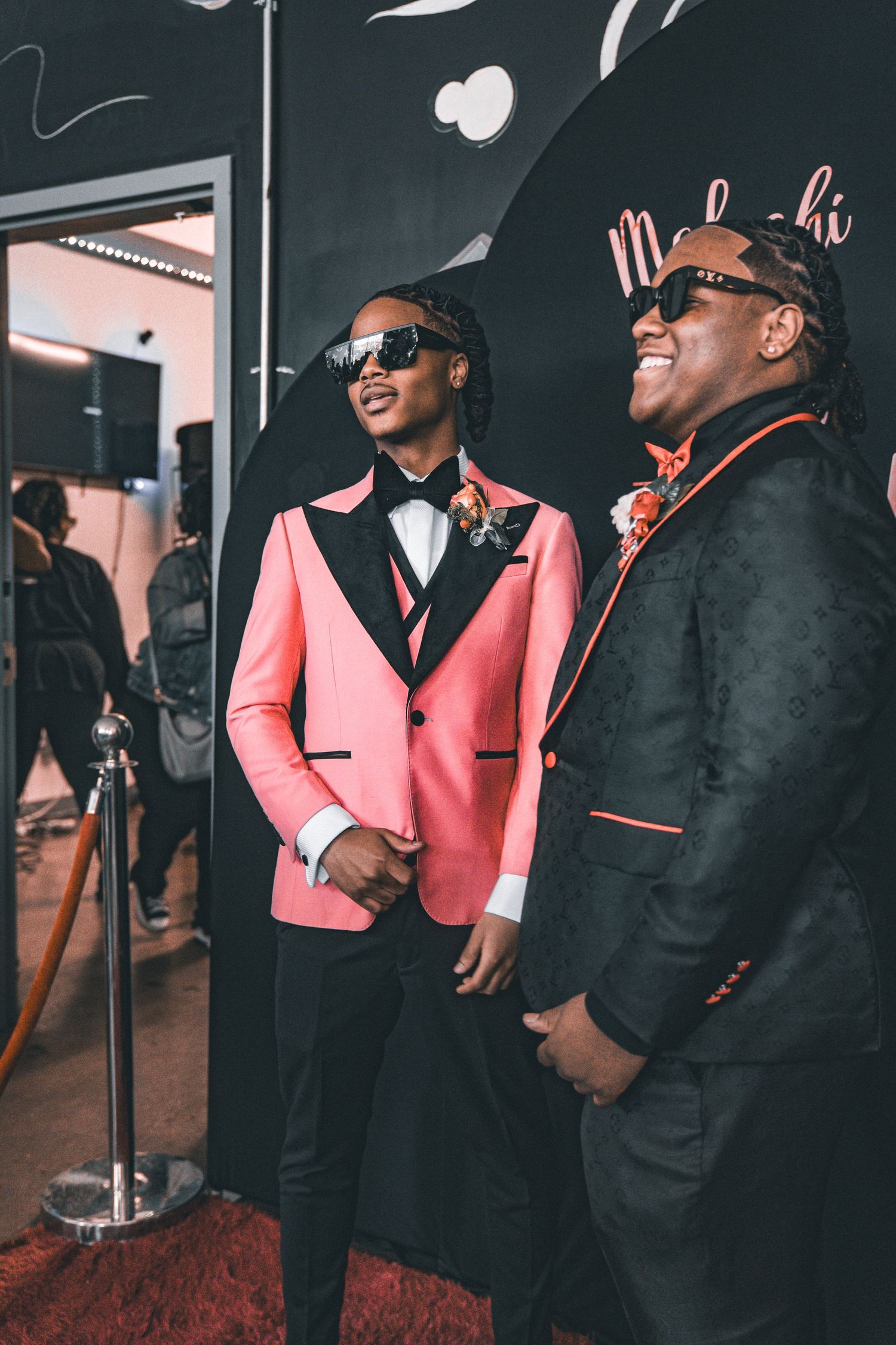 Two guests wearing formal attire, one in a pink blazer and another in black, share a laugh at an event.