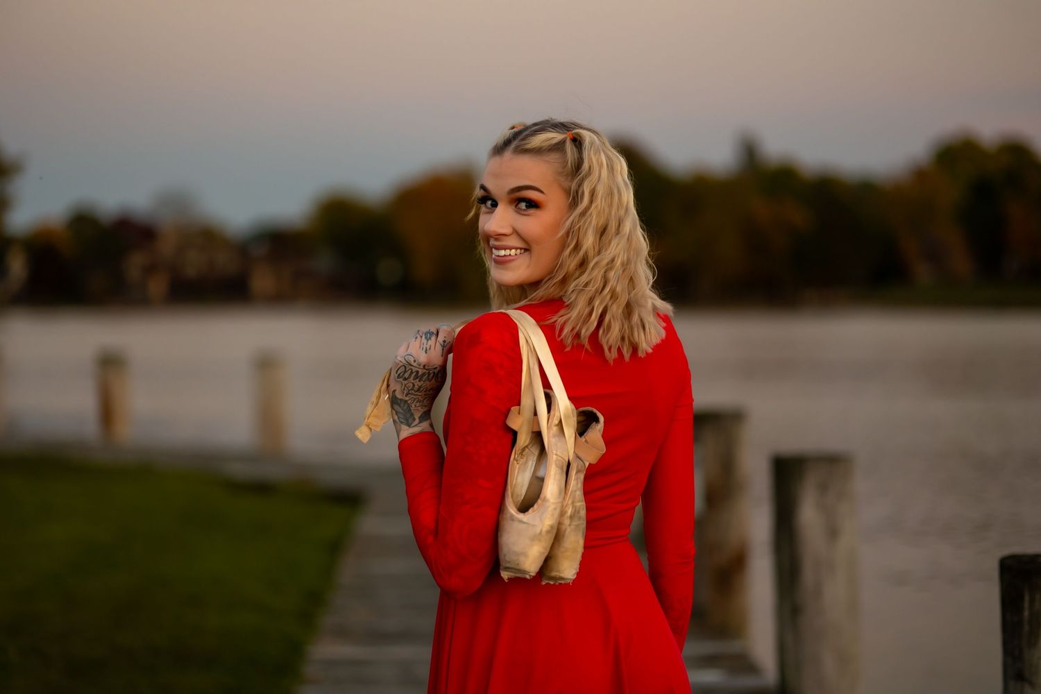 Person in red dress holding ballet pointe shoes by lake during sunset.