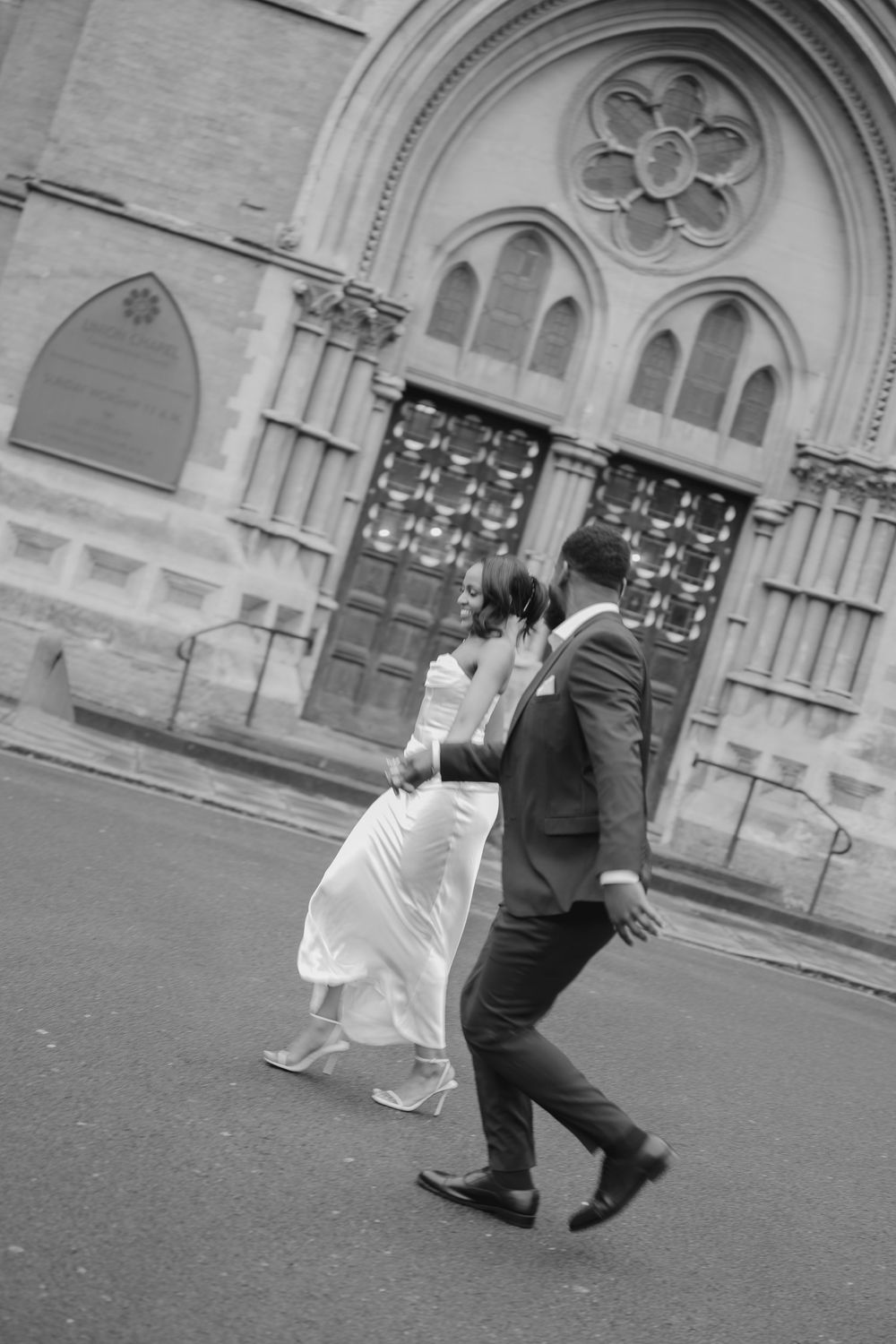 Black and white photo of a bride and groom walking in front of a historic church on their wedding day.