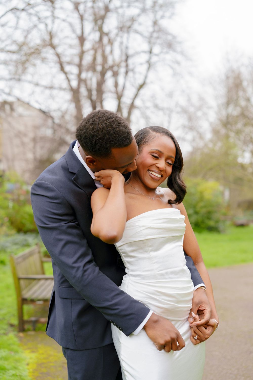 A joyful bride in a white strapless gown laughs as her groom in a navy suit embraces her in a lush green park.