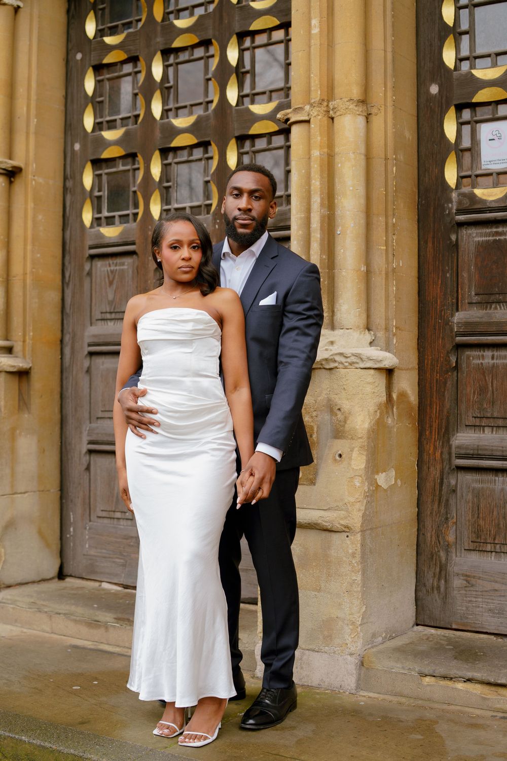 Elegant couple posing in front of ornate wooden doors, woman in white satin gown, man in gray suit holding hands.
