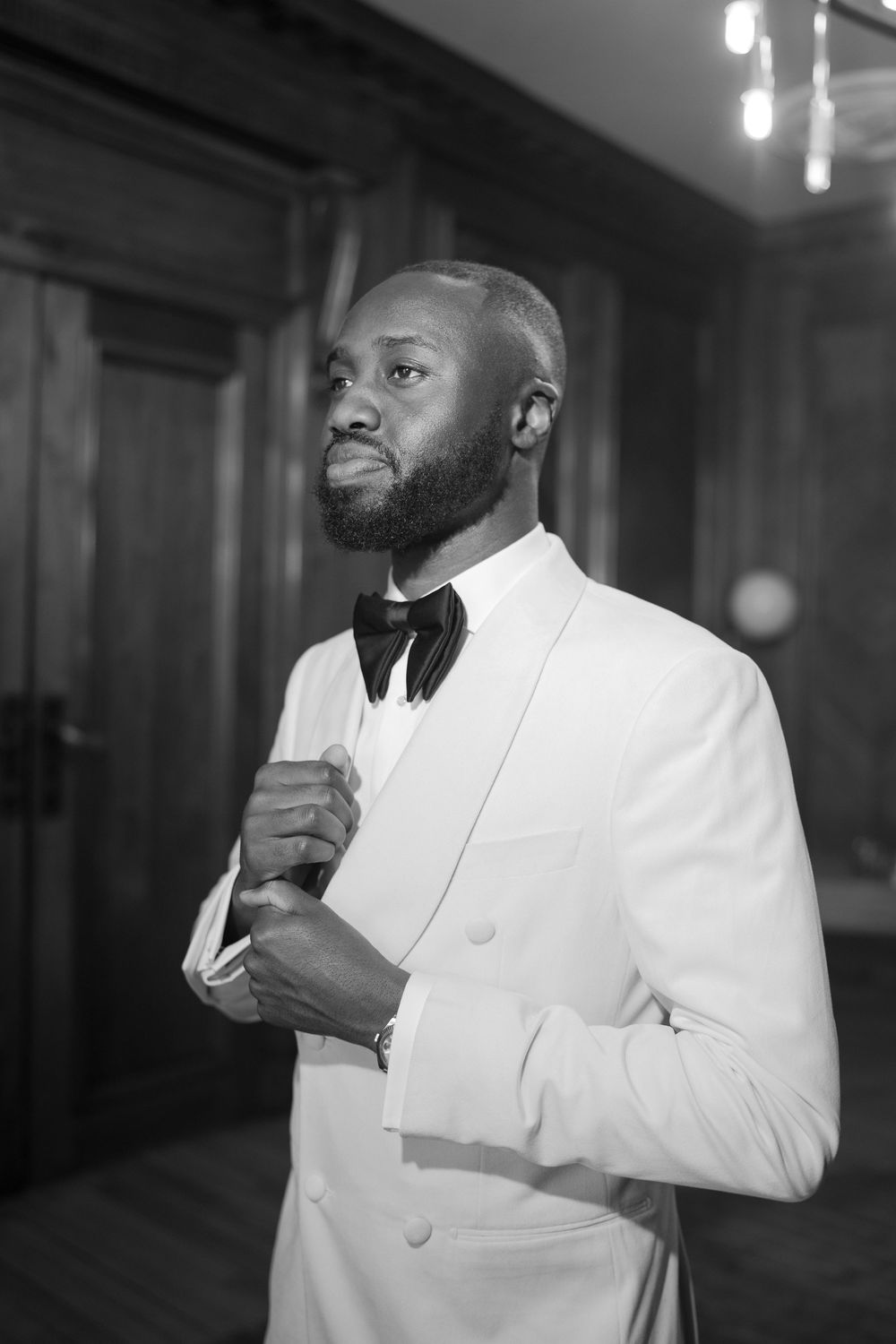 A black and white photo of someone wearing a white tuxedo jacket and black bowtie adjusting their collar.