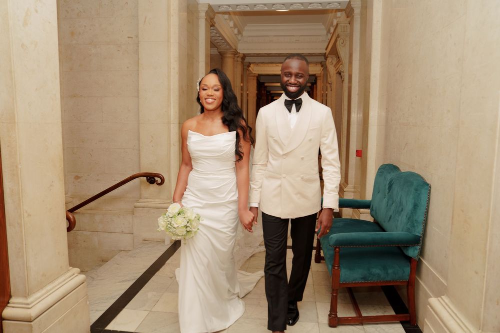 A couple walking together in a formal hallway, the bride in white gown holding flowers and groom in cream tuxedo.