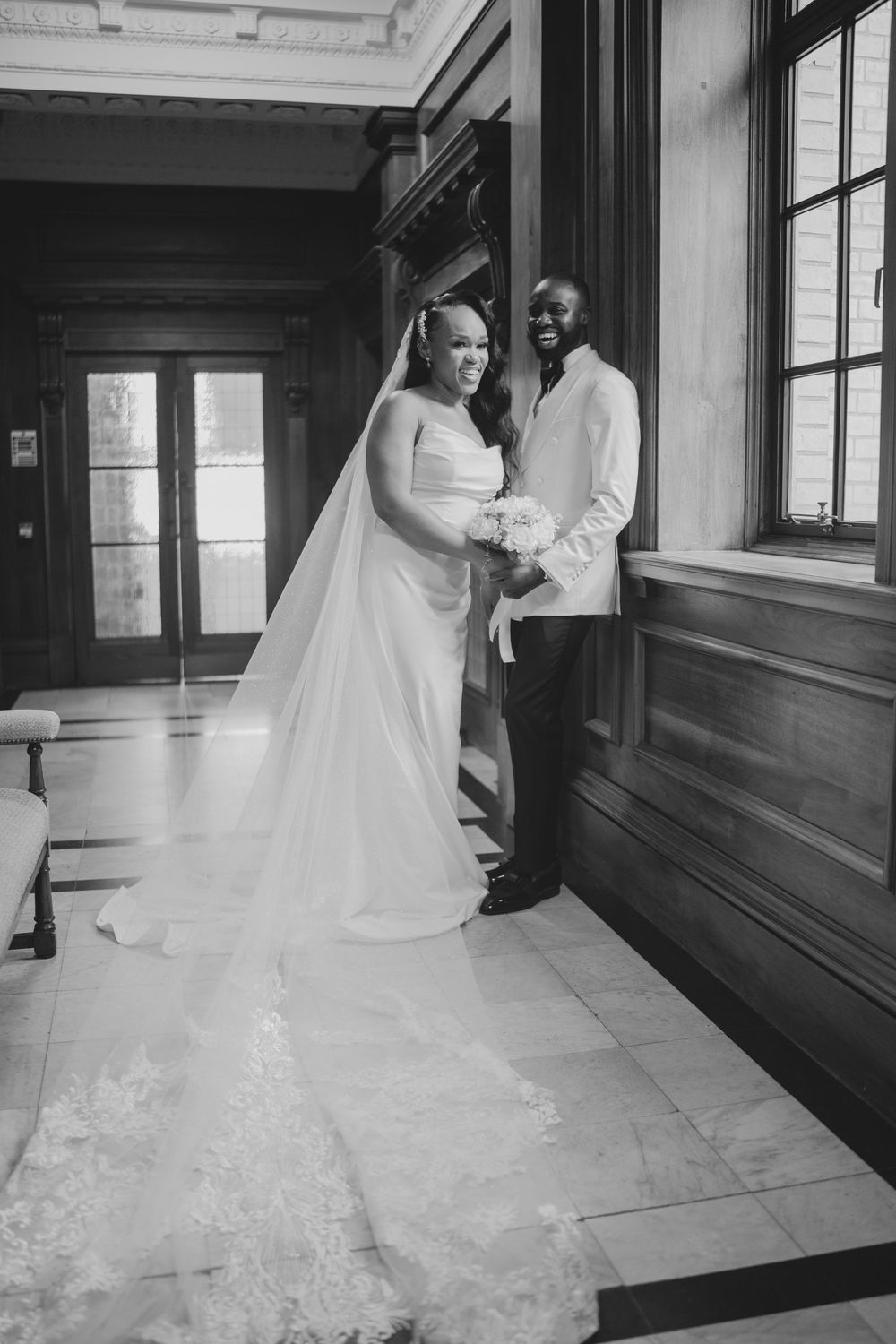 A bride in an elegant white gown and veil stands in a historic building interior with wood paneled walls.