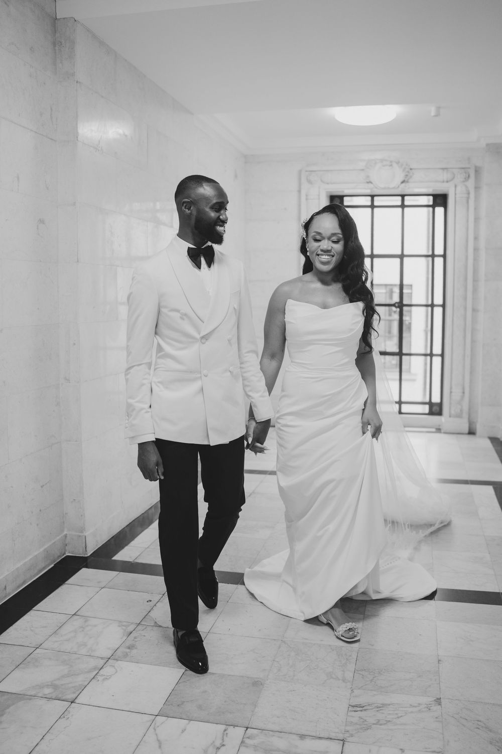 A black and white wedding photo of a couple walking hand in hand down a hallway, wearing elegant formal attire.