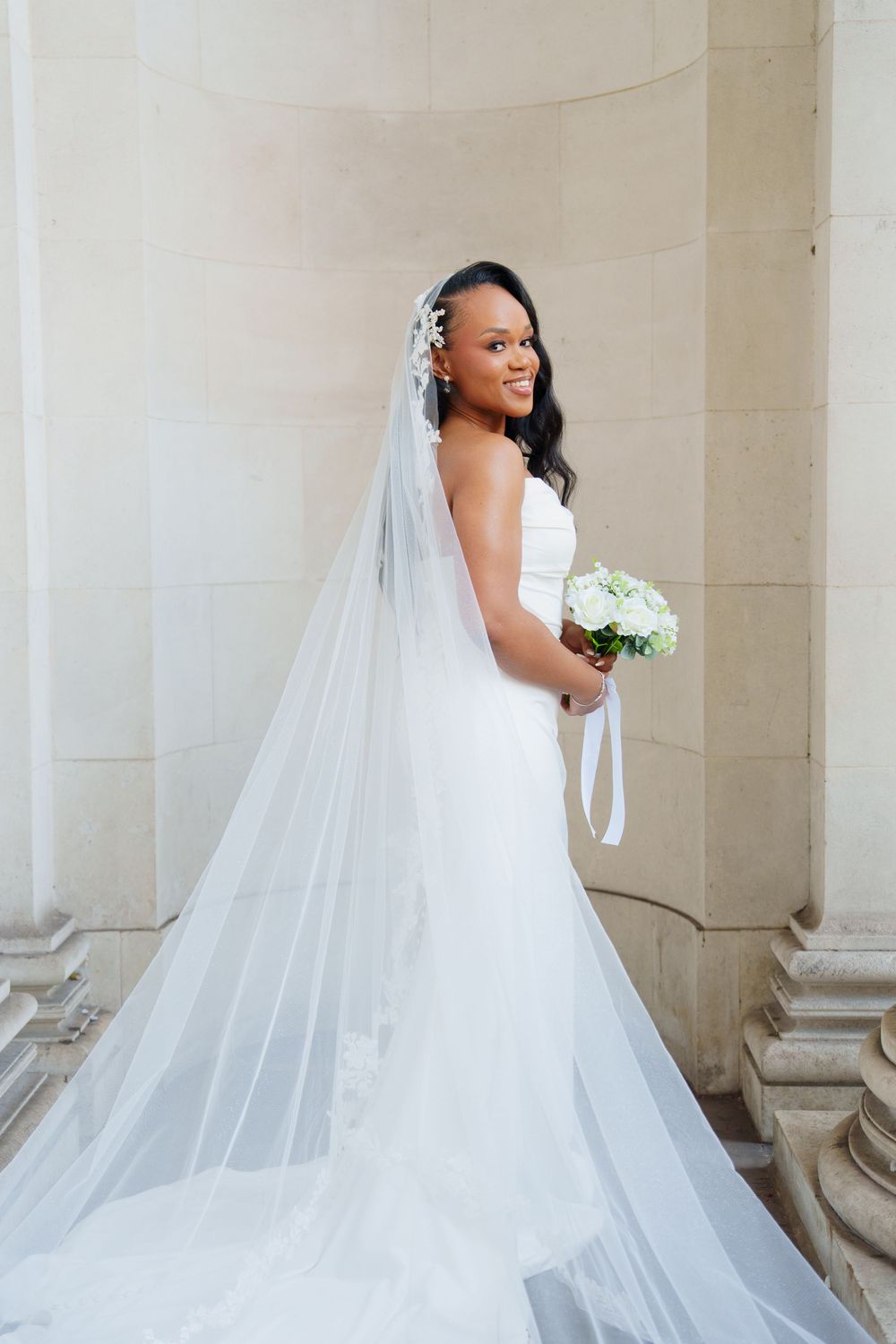 A bride in an elegant white wedding gown with a flowing cathedral veil poses gracefully against a light stone wall.