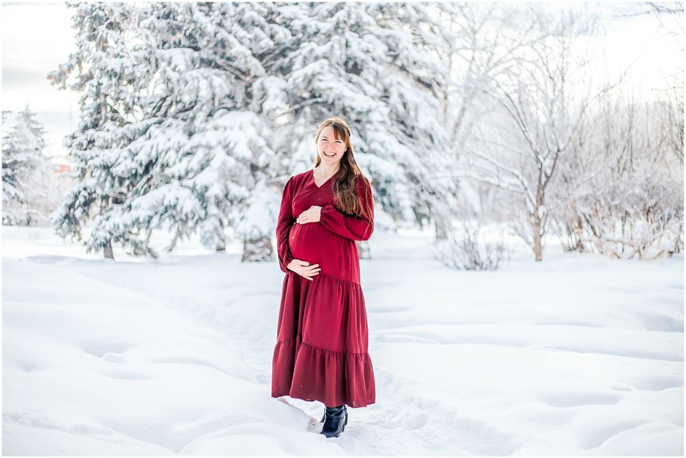 Someone in a flowing burgundy dress stands in a serene winter landscape with snow-covered evergreen trees.