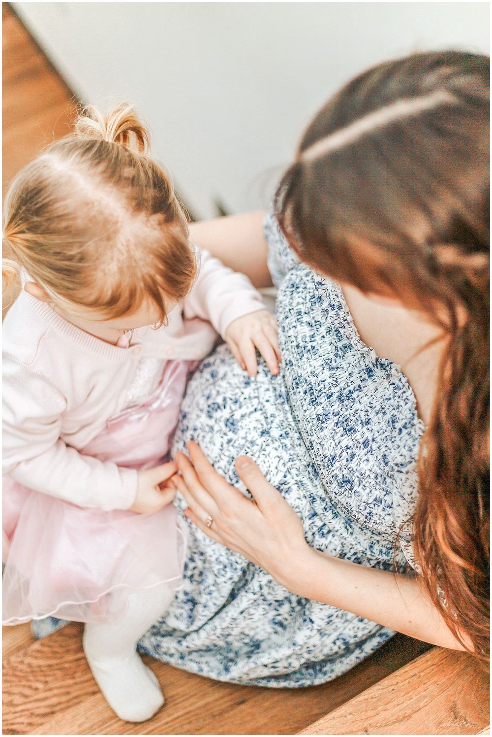 A tender moment between mother and daughter on a hardwood floor, both wearing floral dresses in pink and blue.