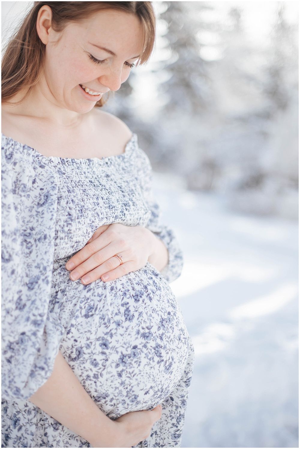Expectant mother in blue floral dress cradles baby bump against snowy winter backdrop in soft natural lighting.