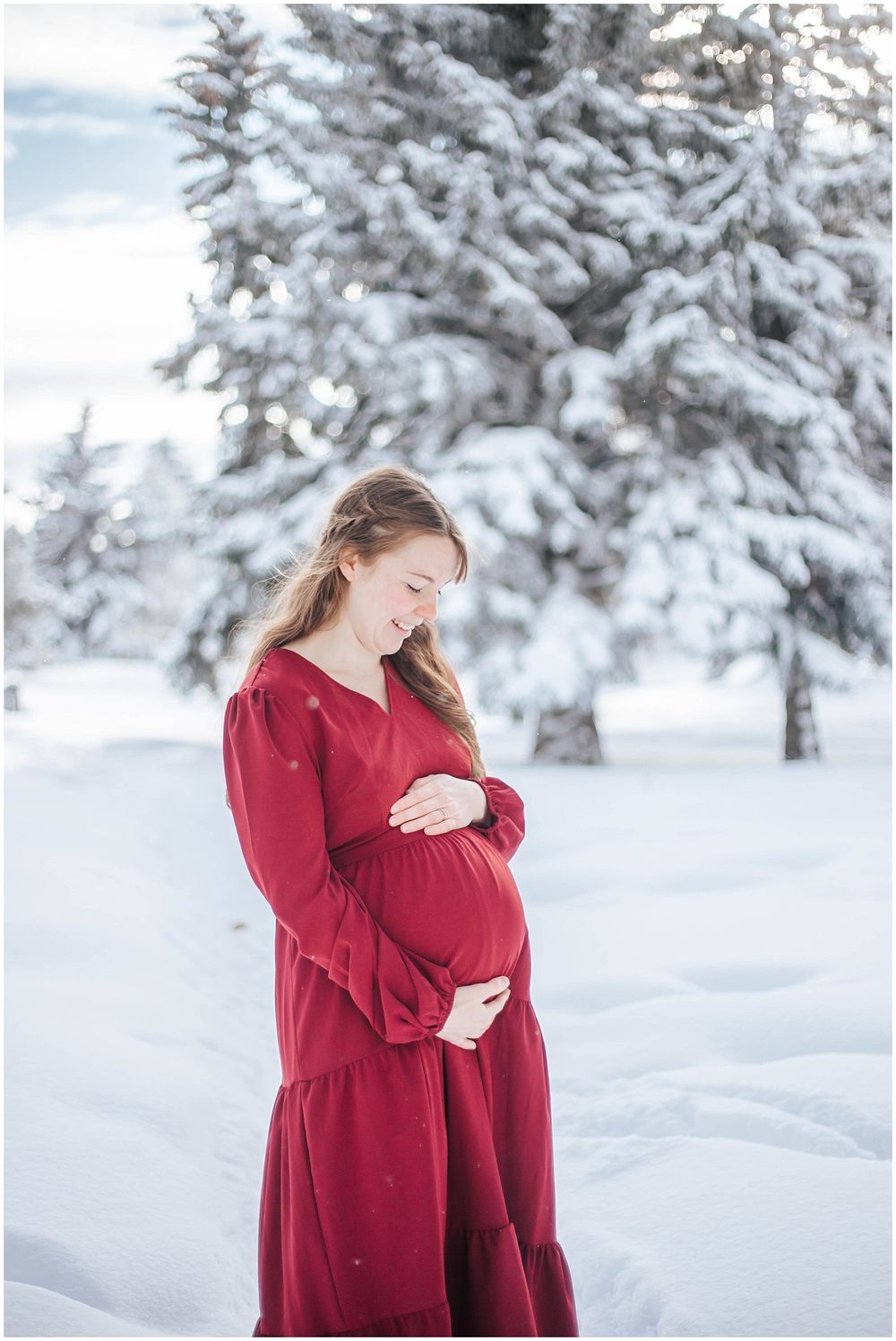 Maternity portrait in winter with snow-covered evergreens and a flowing red dress against white snow.