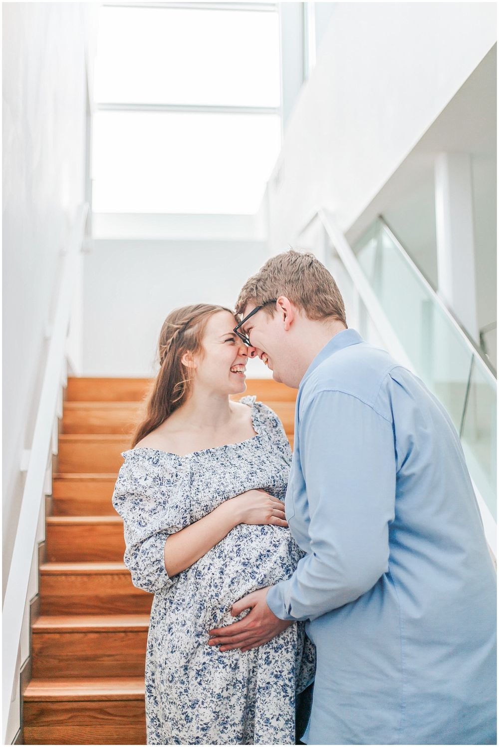 A couple in casual attire share an intimate moment on wooden stairs near a bright window.