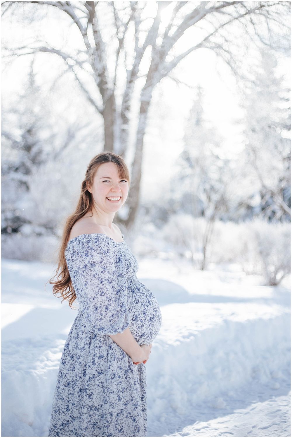 A woman in a blue floral dress poses for a maternity photo in a snowy winter landscape.