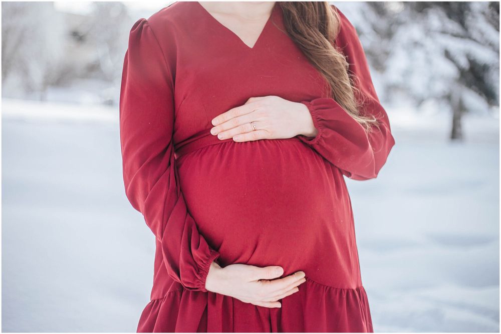 Someone in a flowing red maternity dress cradles their pregnant belly while standing in a snowy winter landscape.
