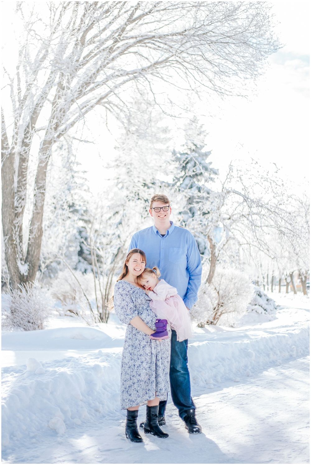 Two people stand together on a snowy winter path surrounded by frost-covered trees in bright white landscape.