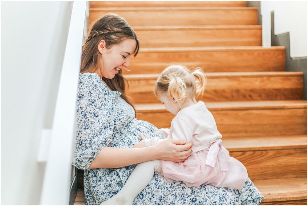 Sweet moment on wooden stairs as mother and daughter share quiet time together in floral dress and pink outfit.