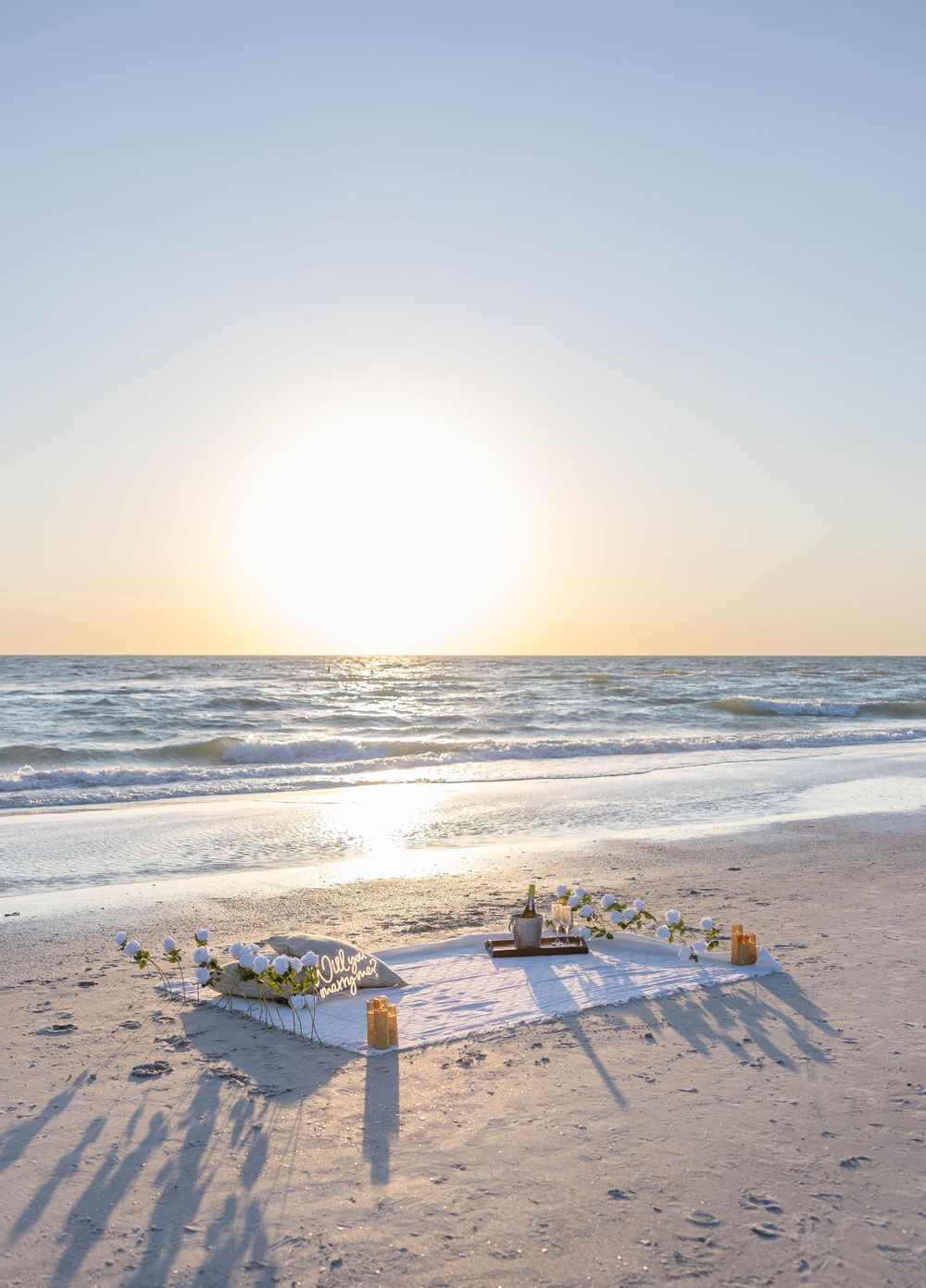 A romantic beach picnic setup with white flowers and candles on the sand at golden sunset by the ocean.