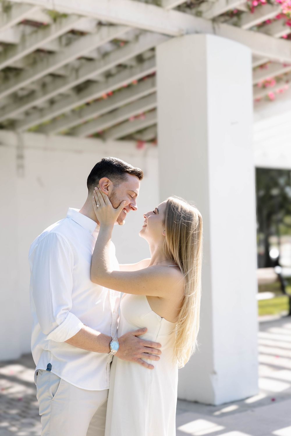 A couple in white outfits share a romantic moment, smiling and embracing under a sunlit pergola with floral accents.