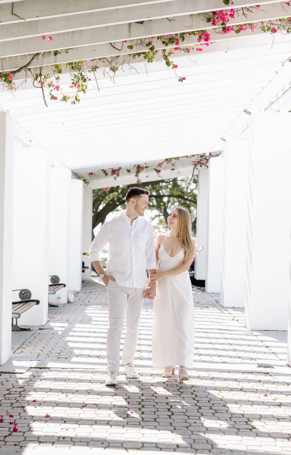 Couple in white outfits walking hand in hand under a bright pergola adorned with pink flowers.