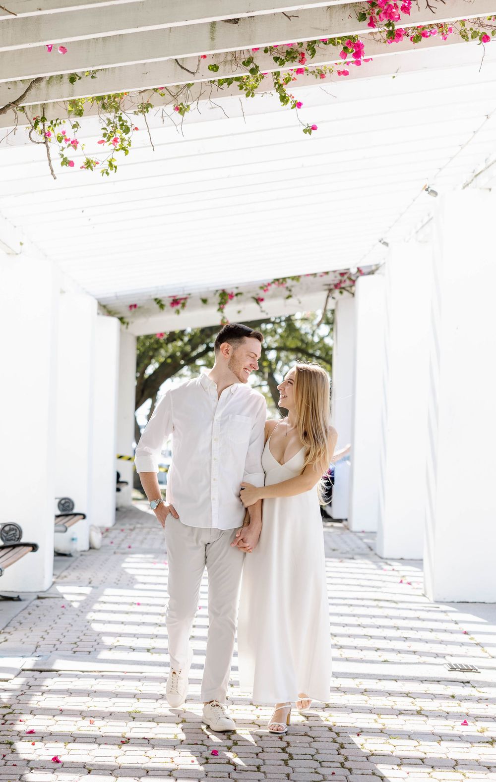 Couple in white outfits smiling at each other under a floral pergola with pink bougainvillea blooms overhead.