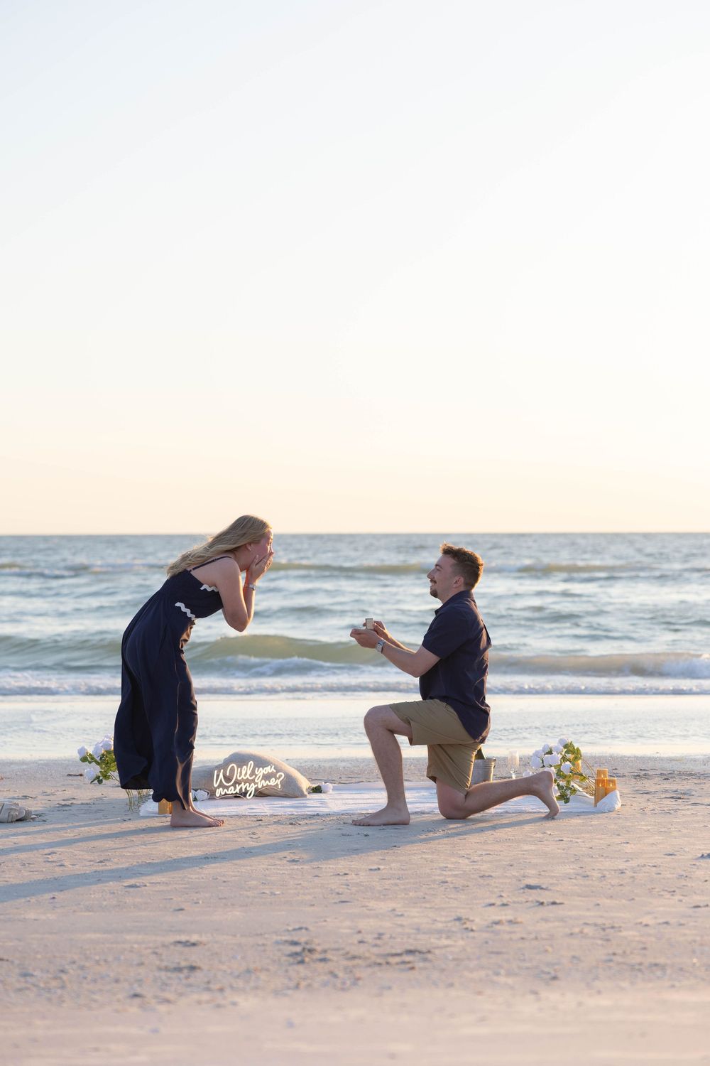 Man proposes on bended knee to woman on sandy beach at sunset with flowers and a sign nearby.