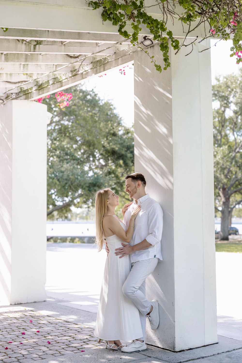 Couple in white outfits sharing a romantic moment under a pergola with lush greenery and bright sunlight.