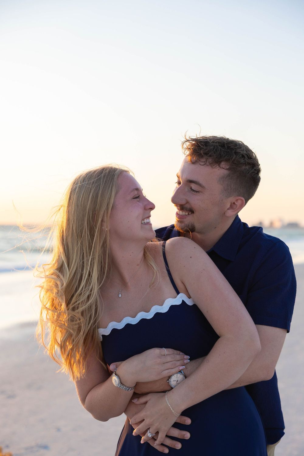 Couple embracing and smiling at each other on a beach at sunset, woman in navy dress, man in navy shirt.