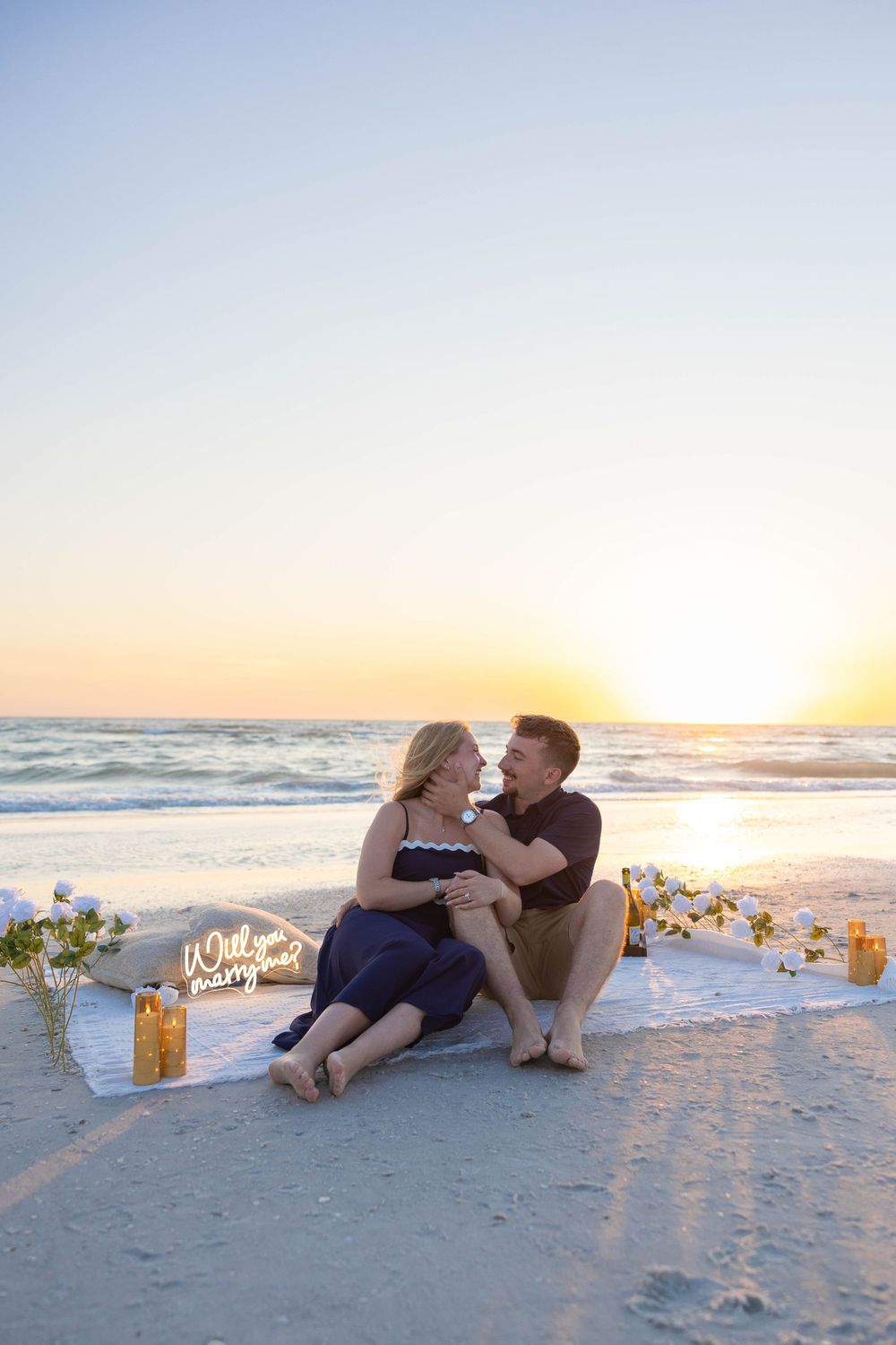 Couple sitting on beach blanket at sunset with Will You Marry Me neon sign during romantic seaside proposal.