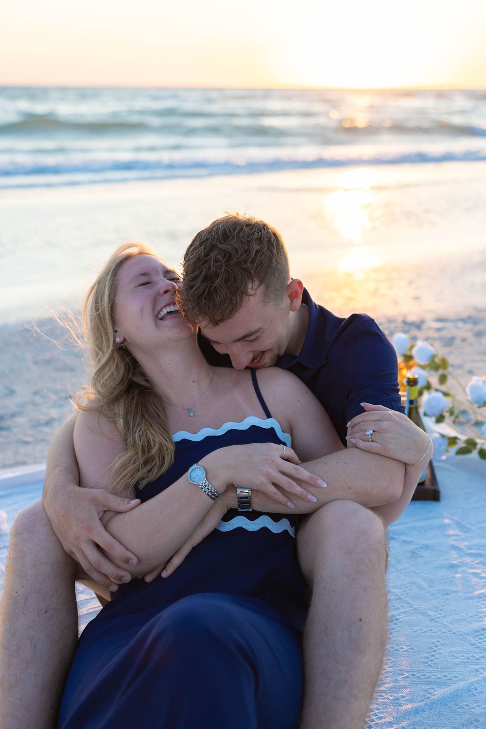 Happy couple embraces and laughs together on beach at golden sunset, woman showing engagement ring.