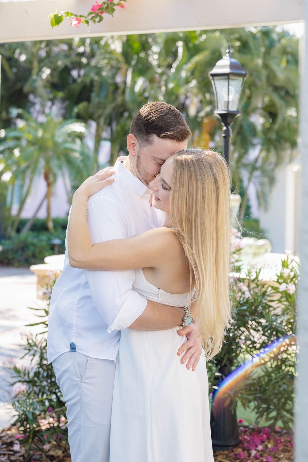 A couple in white outfits embracing and kissing in a tropical garden setting with palm trees and lush greenery.