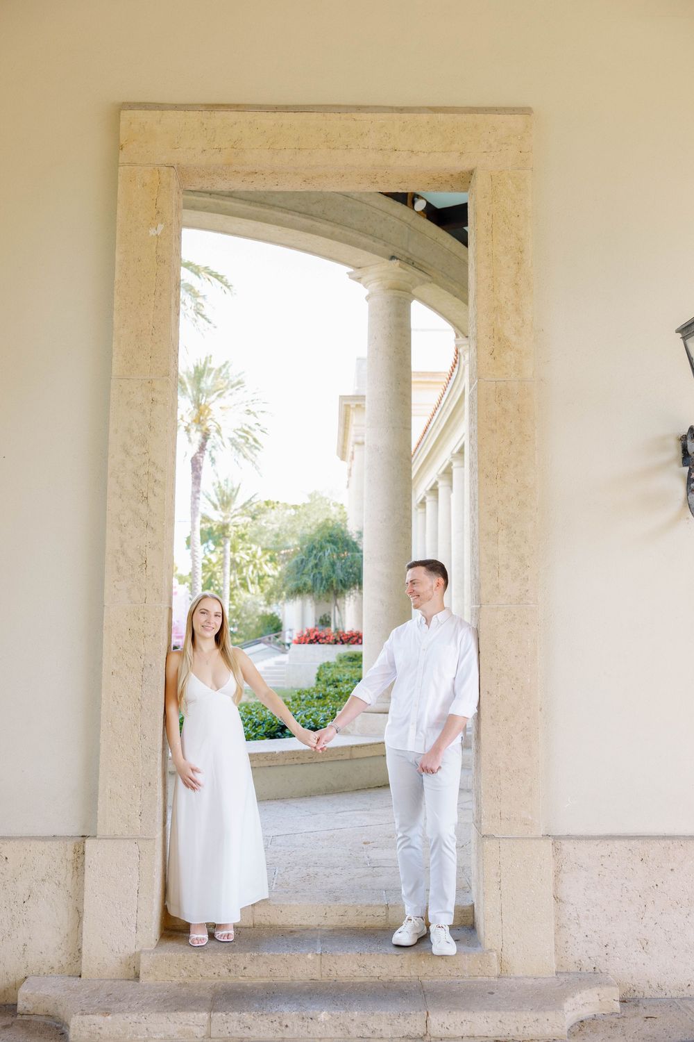 Couple in white outfits holding hands under a stone arch at an elegant outdoor venue with columns and palm trees.