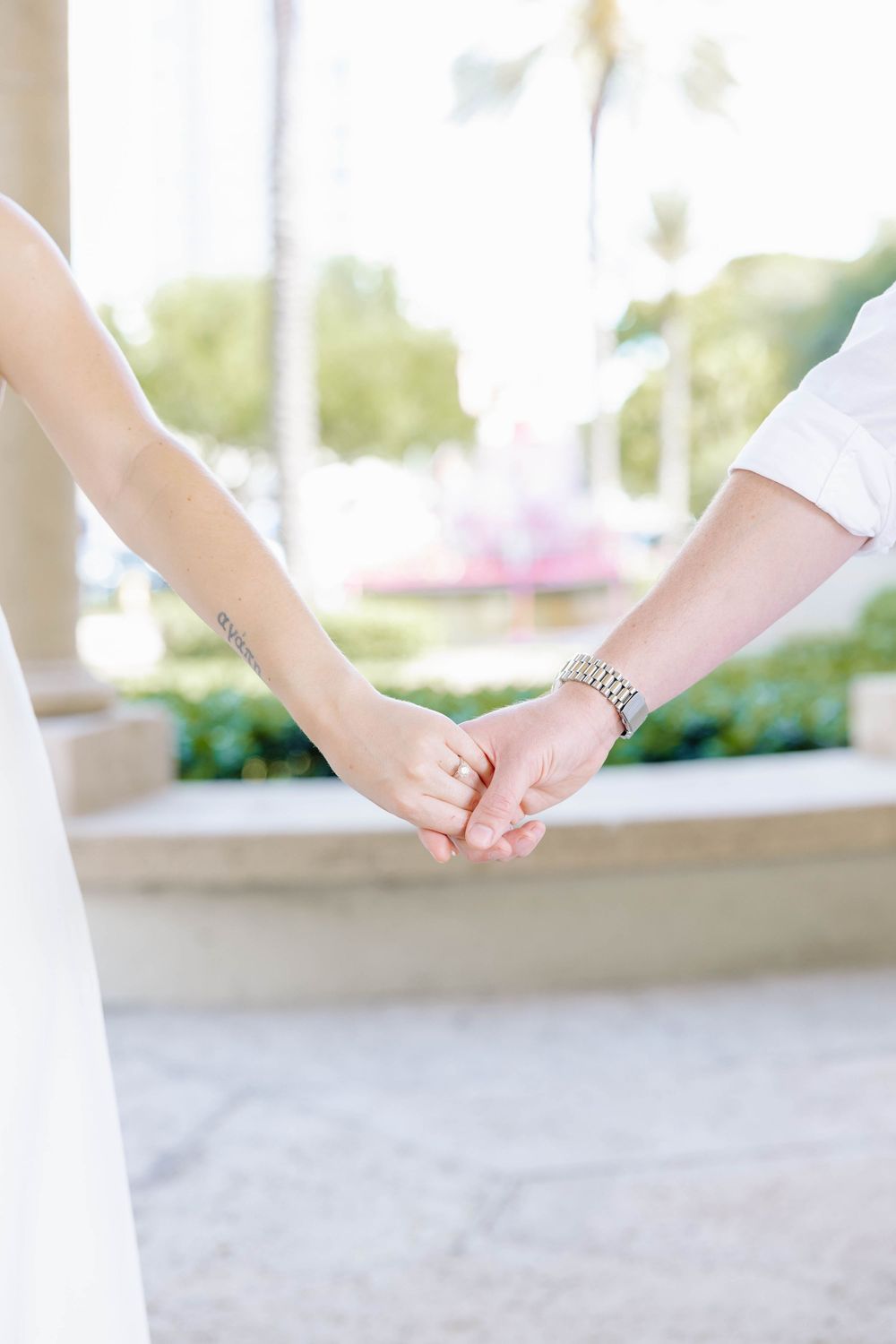 Close-up of a couple holding hands, bride wearing engagement ring, groom with silver bracelet, outdoor garden background.