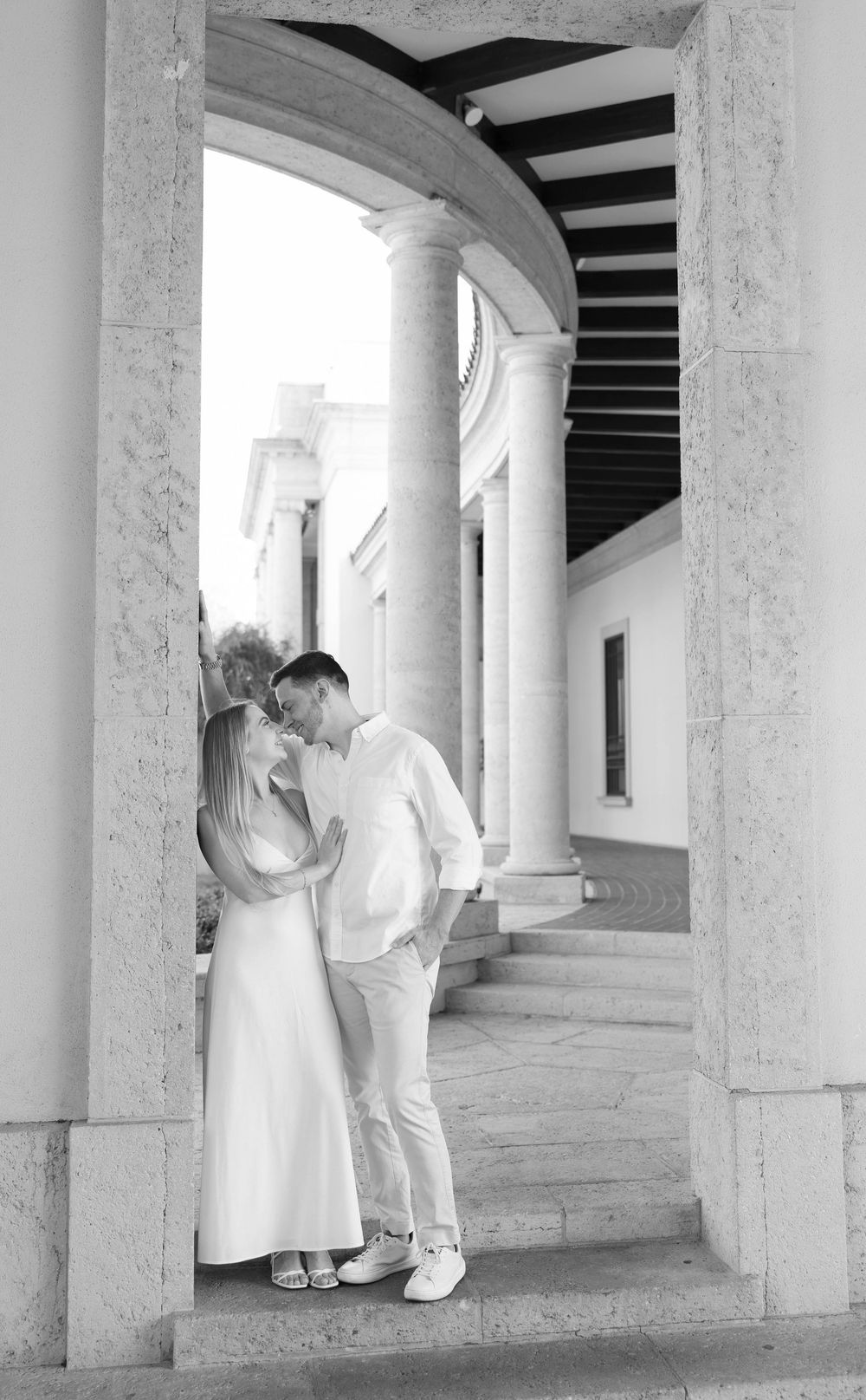 Black and white photo of a couple in white outfits sharing a romantic moment under elegant stone archway columns.