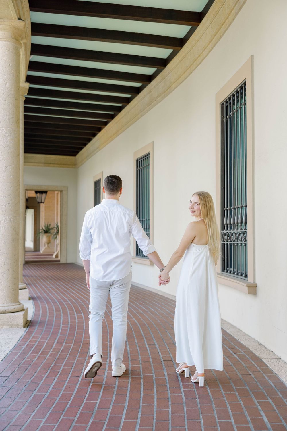 Couple in white outfits holding hands while walking through an elegant outdoor corridor with brick flooring.