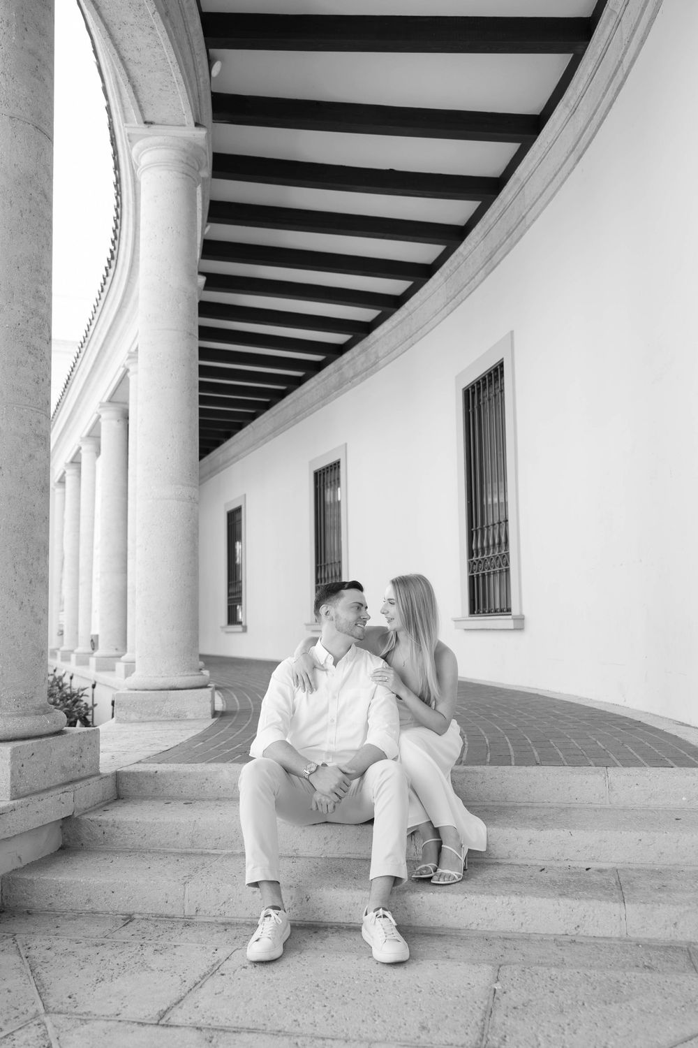 Black and white photo of a couple sitting on steps under elegant columns, sharing a romantic moment.