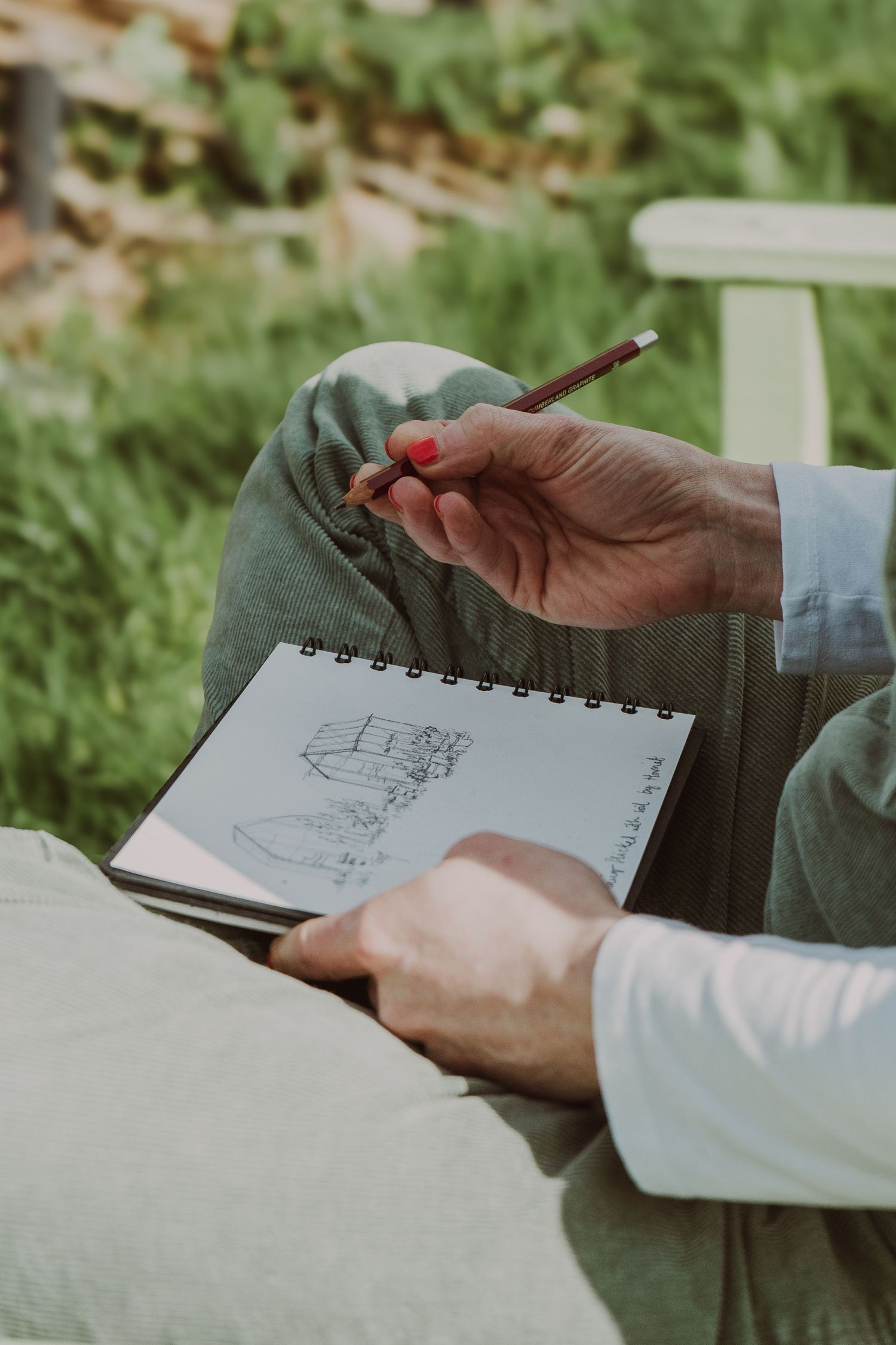 a close up photograph from a branding photo session of a white female in an allotment, sitting on a bench whilst drawing in a sketch book
