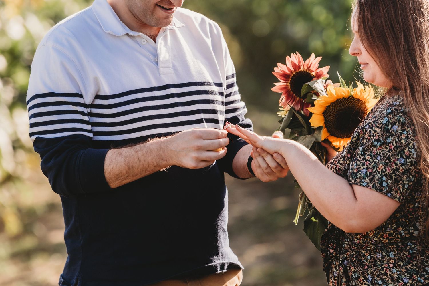 Two people's hands exchanging a small item as they hold vibrant sunflowers on a sunny day outdoors.
