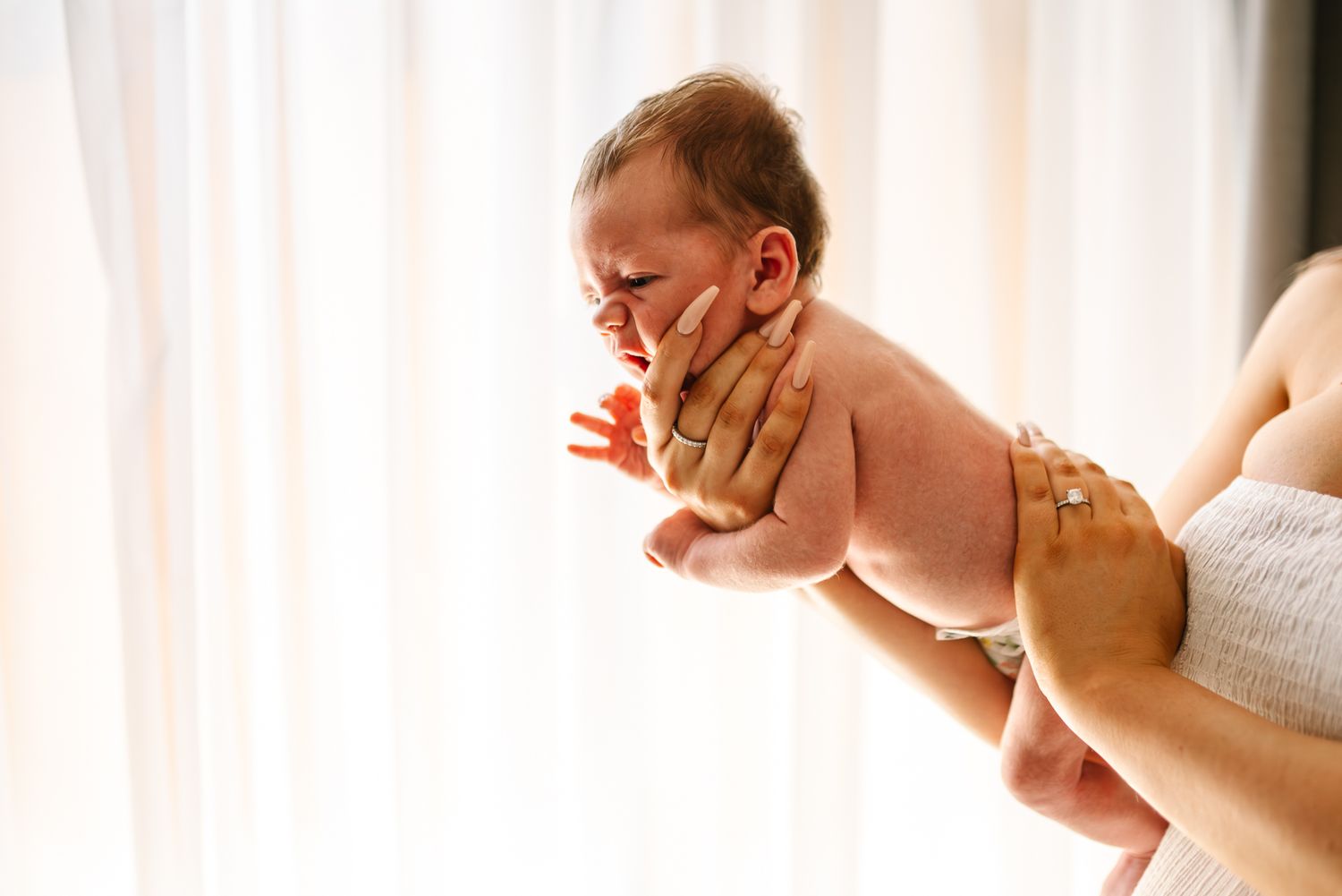 Baby in a white diaper being lifted up against sunlit window curtains during taken by Melbourne Newborn photographer.