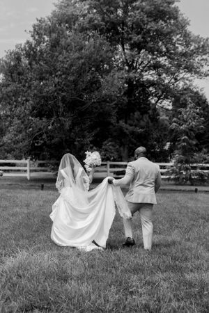 Black and white photo of newlyweds dancing playfully in a grassy field with trees in background.