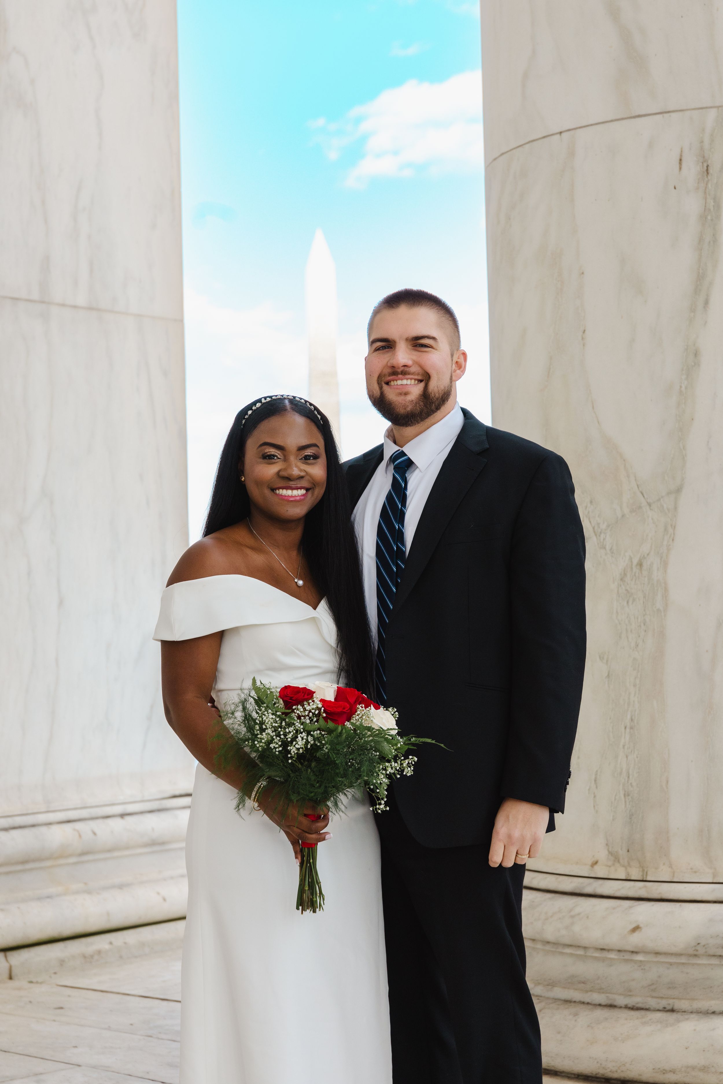 Newlyweds pose together with red bouquet against marble columns on their wedding day.