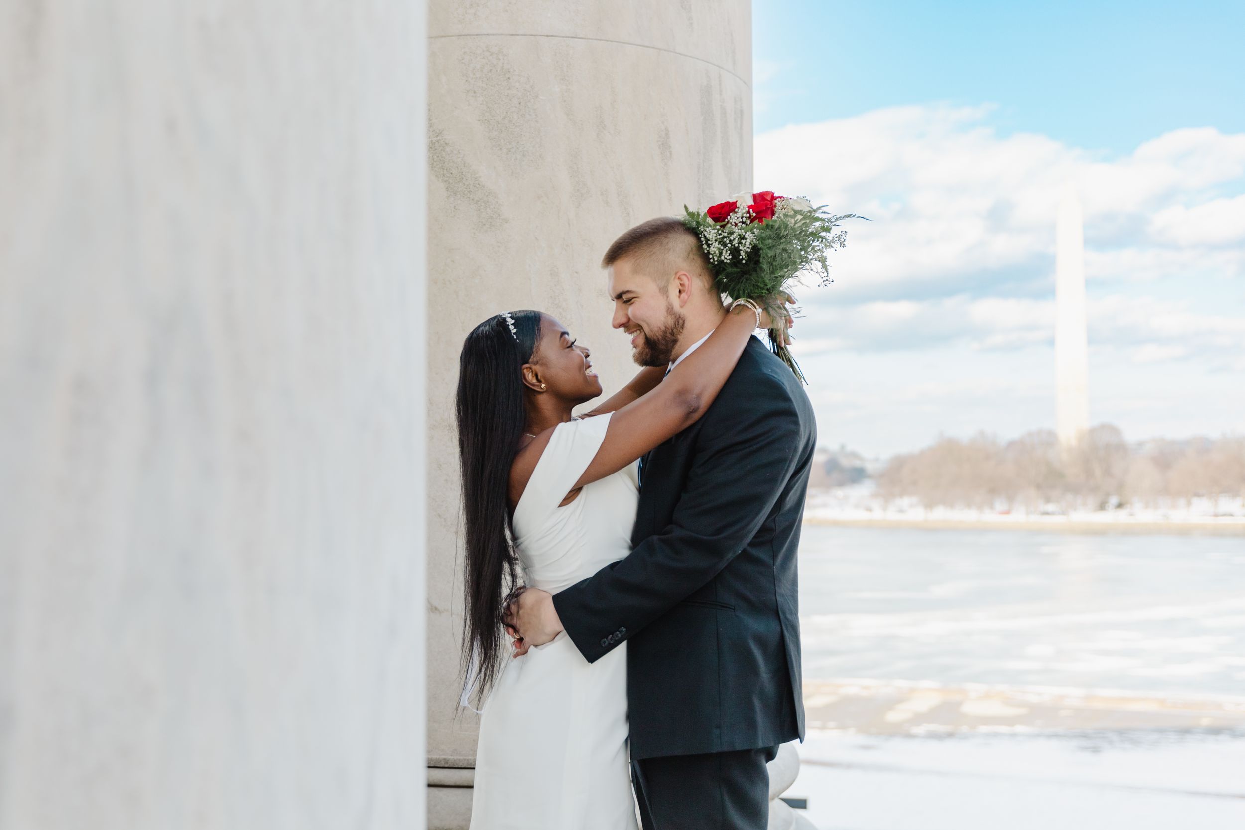 Wedding couple embraces between classical white marble columns on a sunny day.
