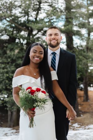 Winter wedding portraits of couple with red bouquet against snowy forest backdrop.